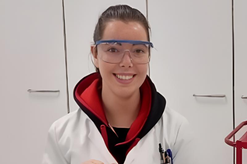 A young woman smiling and standing in a laboratory setting with white cabinets and laboratory equipment in the background. Pens are visible in the pocket of her lab coat.